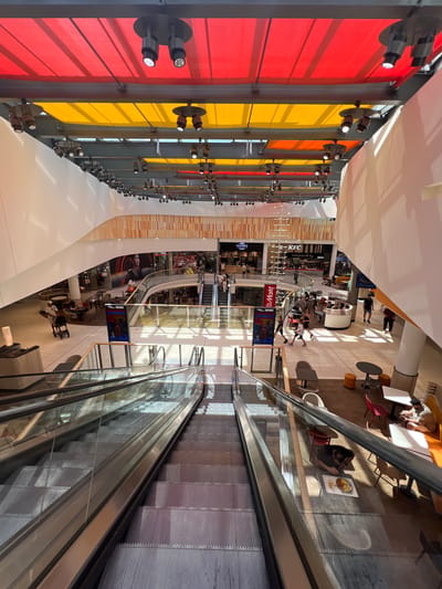 The image captures the interior of a modern shopping mall, likely in Aachen, Germany, characterized by its expansive, multi-level design and vibrant overhead lighting. The upper ceiling features alternating panels of red and yellow fabric, interspersed with a network of spotlights, creating a dynamic visual effect. Below this, the mall's interior reveals multiple levels with shops and common areas.
In the foreground, a well-lit escalator descends, its metallic rails and glass panels reflecting the ambient light. Several people are visible in the midground and background, navigating the mall. On the ground floor, some individuals are seated at tables in a dining area, while others walk through the open spaces between shops.
Notable elements include storefronts with large displays, such as one for "Yamens" and another for "KFC." Digital screens are also present, one displaying "NEWS." The architecture is contemporary, with curved walls and sleek finishes. Sunlight streams in from unseen openings, casting shadows and highlighting the polished floors. The overall impression is one of a busy, well-maintained commercial environment.