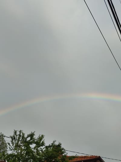 This media file captures a sky with a faint rainbow arching across it. The sky is overcast with greyish clouds, suggesting a recent or ongoing rain shower. In the upper left quadrant, dark power lines cut diagonally across the frame. To the right, a portion of a building with a red tiled roof and a brick facade is visible, along with the branches of a lush green tree. The presence of power lines and a building indicates an urban or suburban setting. The time of day appears to be daytime, given the ambient light. The overall mood evoked is serene, perhaps even hopeful due to the presence of the rainbow, a symbol of beauty after the rain. There is no visible text in the image. The location context of Pletena, Bulgaria, places this scene in Eastern Europe.