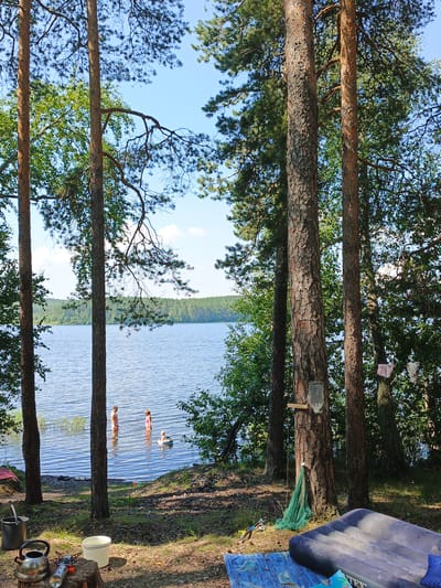 This is a medium-shot, eye-level, outdoor, vertical photograph taken at a lake in Nepryakhinskoye Sel'skoye Poseleniye, Russia. In the foreground, there are several objects on the ground including a camping stove, a kettle, a bucket, and an inflatable mattress. In the middle ground, three people are in the water, two standing and one on a float. The background features a body of water and a dense forest. The lighting is natural and bright, suggesting it is daytime. The overall scene depicts a recreational setting at a lake.