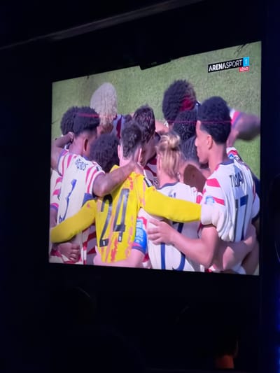 The image shows a screen displaying a sports broadcast. The broadcast features a group of soccer players in a huddle, wearing white jerseys with red and blue accents. One player in the foreground is wearing a yellow jersey with the number 24 on the back. The broadcast logo in the upper right corner reads "ARENASPORT 1 UŽIVO". The screen is positioned in a dark room, likely a home or bar, with the bottom of the screen obscured by darkness. There is no visible information in the image that would place it in Travnik, Bosnia and Herzegovina.