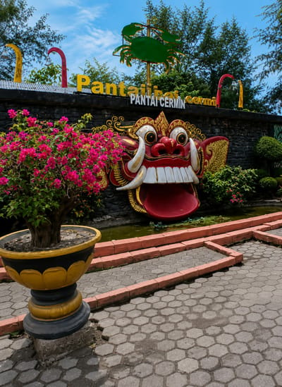 In the city of Kota Pari, Indonesia, a vibrant and slightly unsettling scene unfolds. Dominating the center of the image is a large, grotesque mask, painted in vivid reds, yellows, and whites. Its wide, toothy grin and bulging eyes give it a fierce, almost monstrous appearance. The mask is mounted on a dark, stone-like wall, which serves as a backdrop. Above the mask, yellow lettering spells out "Pantai" and "Pantai Cermin" in Indonesian, indicating the location is a beach called Cermin Beach. Flanking the mask on the wall are decorative elements, including stylized golden wings and red and yellow poles. In front of the wall, a small pond with green water is visible, bordered by coral-colored brick steps. To the left of the mask, a potted plant with abundant pink bougainvillea flowers adds a splash of color and softness to the otherwise intense imagery. The plant sits in a large, decorative black and yellow planter. The foreground is paved with hexagonal grey stones. The sky above is a clear blue with scattered white clouds. The overall impression is one of a tropical, possibly tourist-oriented location with a unique, culturally inspired artistic expression.