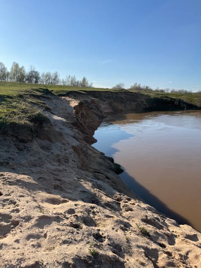 The image shows a view of a steep, sandy embankment overlooking a body of water, likely a river or a large pond, and its surroundings. The embankment is light in color, suggesting sand or loess soil. The water is brownish, indicating sediment content. Vegetation, including grass and bare trees, grows at the top of the embankment away from the water. The sky is clear blue. Based on the user's request of the location being Voronino, Russia, it can be inferred that this is a landscape found near Voronino, Russia, possibly along a riverbank.