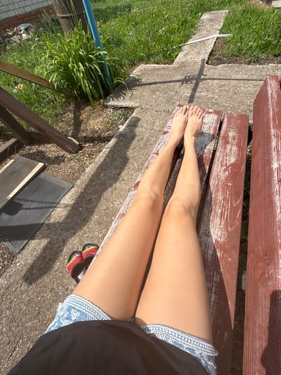 A first-person perspective shot shows a person's legs and feet resting on a weathered, red-brown wooden bench. The person is wearing denim shorts and black sandals with colorful straps. The legs are tanned and appear to be bathed in sunlight, casting a shadow on the concrete ground. The surroundings suggest an outdoor setting, possibly a yard or garden, with green grass, a concrete pathway, and some foliage. A partial view of a brick wall and wooden fencing is visible in the background. The lighting indicates it is daytime, with the sun casting strong shadows.