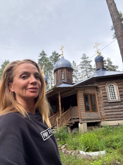 The image shows a woman with blonde hair taking a selfie in front of a wooden Russian Orthodox church. The church has a dark blue dome topped with a golden cross. The woman is wearing a black sweatshirt with white text that appears to read "PURPP TH". She has hoop earrings and is smiling. The church is surrounded by trees and greenery. The scene appears to be located in a rural area, possibly in the village of Nenimäki, Russia, given the architectural style of the church and the surrounding natural environment. The sky is overcast.