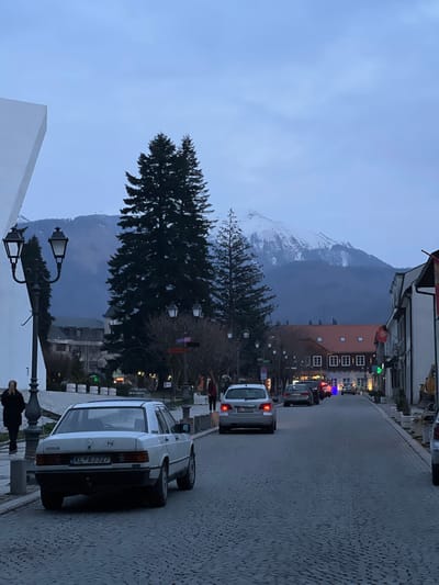 The image depicts a street scene in the city of Kolašin, Montenegro, at twilight or dawn. A cobblestone street recedes into the background, flanked by buildings and trees.
In the far background, snow-capped mountains are visible under an overcast, blue-grey sky. Tall, dark evergreen trees stand prominently along the street, particularly on the left side, obscuring parts of the distant landscape.
On the left side of the street, a large, angular white architectural structure is partially visible. Two ornate black streetlights with multiple lanterns are positioned in the left foreground. Two human figures, appearing as silhouettes, walk away from the camera on the left sidewalk.
In the immediate foreground, a white Mercedes-Benz 190D sedan, bearing Montenegrin license plate KL AJ327, is parked on the left side of the cobblestone street, facing away from the camera. It is equipped with a tow hitch. Further down the street, a silver station wagon or hatchback with illuminated taillights, bearing Montenegrin license plate KL BQ061, is moving away from the camera. Several other vehicles, some with illuminated taillights, are visible further down the street.
The right side of the street is lined with multi-story buildings featuring traditional architecture, including pitched roofs, some with red tiles. Commercial establishments are indicated by various signs, including a green cross symbol visible on one building. One human figure in dark clothing is walking away from the camera on the right sidewalk. Streetlights line both sides of the street, some appearing unlit.