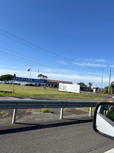 A roadside view shows a blue sign reading "Panorama House," with "Restaurant Function Centre Bar" beneath, next to two flags on poles. A low, modern building is set back in a grassy area. A metal guardrail runs along the edge of the road, and the side mirror of a car is visible in the upper right. Power lines stretch across a clear blue sky with faint clouds.