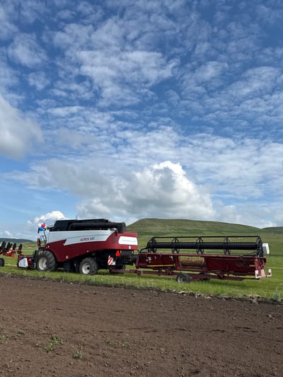 A combine harvester and its header attachment are parked on a field in Старое Субханкулово, Russia. The combine is red and white with "ACROS 585" written on its side. The header, also red, is attached to the front of the combine. The field is brown and appears to be recently tilled. In the background, there are green hills under a blue sky with scattered white clouds. The scene suggests agricultural activity.