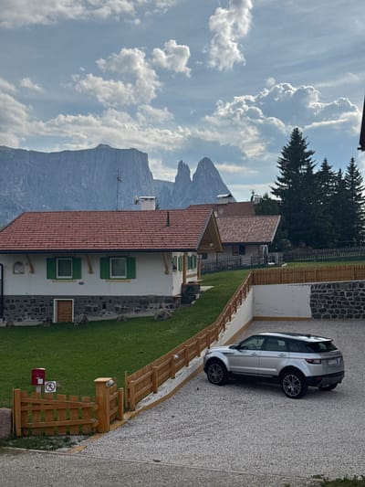 The image captures a scenic view of the Dolomites in Kastelruth (Castelrotto), Italy. The scene is dominated by a silver Range Rover parked on a gravel driveway next to a modern building with a terracotta tiled roof and white walls. The building features green-shuttered windows. In the background, dramatic mountain peaks rise against a sky filled with fluffy white clouds. Tall evergreen trees stand to the right of the frame, casting shadows on the landscape. A wooden fence lines the edge of the lawn, with a red mailbox and a circular sign depicting a prohibition symbol. The time of day appears to be late afternoon or early evening, given the lighting. The weather seems pleasant with a mix of sun and clouds. The overall impression is one of tranquility and natural beauty, characteristic of the South Tyrolean Alps. The license plate on the Range Rover is partially visible and appears to read "BZ 277EE".