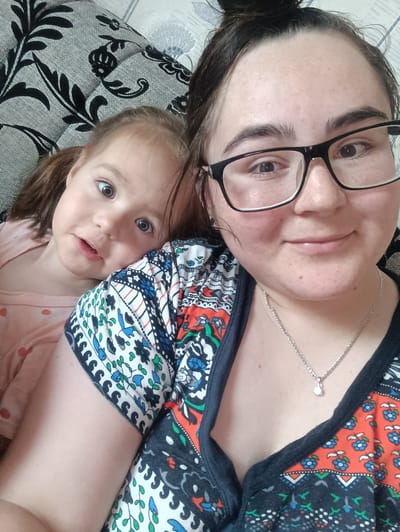 A woman and a young girl are posing for a selfie on a couch. The woman is in the foreground, wearing glasses and a colorful shirt. Her hair is pulled up in a bun. The girl is in the background, leaning on the woman's shoulder. She has her hair in pigtails and is wearing a pink shirt with red polka dots. The couch has a black and white patterned fabric. The lighting suggests it's daytime. The scene appears to be indoors, likely a living room. The overall mood is relaxed and affectionate. No visible text is present.