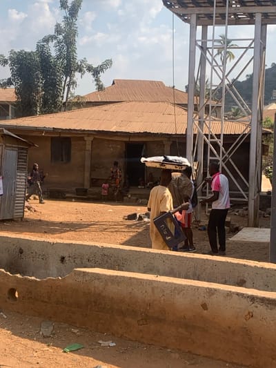 The image depicts an outdoor scene in Egbedi, Nigeria, under a bright, partly cloudy sky. The ground is composed of reddish-brown dirt.
In the mid-ground, a single-story building with light-colored, possibly stucco or mud walls and a rust-colored corrugated metal roof dominates the left and center. It features multiple windows, some with visible shutters, and a central doorway where several individuals are gathered. One woman in a colorful patterned dress and two children in pink clothing are discernible near this entrance. To the far left of this building, a smaller, corrugated metal shack is visible.
To the right of the main building, a tall, grey metal lattice tower stands with a wooden platform at its apex. A thin cable or wire extends downwards from the tower's structure. In the foreground, two parallel, weathered concrete barriers or channels run horizontally across the lower portion of the frame. Scattered debris, including green plastic and white paper, is visible on the dirt ground between and around these barriers.
Several individuals are present. In the right-center mid-ground, a man in a light-colored, long traditional garment (possibly an agbada) holds a blue plastic crate. Adjacent to him, another man balances a white tray containing dark objects on his head, wearing a dark shirt and shorts. A third man in a white and pink polo shirt and dark pants stands facing these two, appearing to interact with them. Further left, in front of the main building, a person in dark, possibly camouflage-patterned clothing with a facial covering walks towards the left, away from the building. In the background, beyond the main building, more structures with similar corrugated roofs are visible, along with a hillside featuring sparse vegetation including palm trees. A large tree with green foliage stands to the left of the main building.