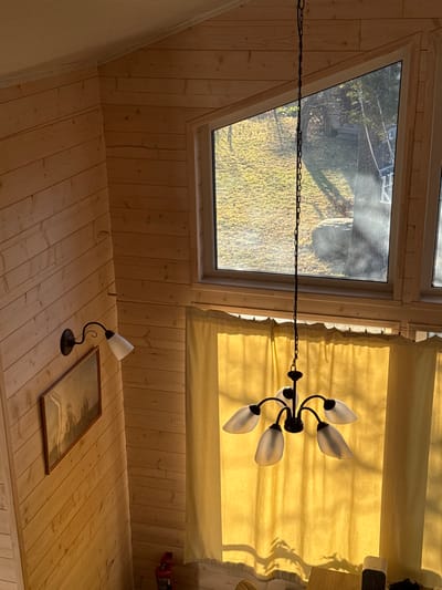 An interior shot from an elevated perspective in Dubki, Russia, captures a room with light-colored wooden plank walls. On the left wall, a dark metal wall sconce with a single upward-facing white frosted glass shade is mounted above a horizontally framed picture depicting a muted landscape. Overhead, a dark metal chandelier with five downward-facing white frosted glass shades hangs from the ceiling by a chain. Directly beyond the chandelier and below a series of light-framed windows, light-yellow fabric curtains are drawn, illuminated by strong sunlight streaming from outside. Through the upper windows, a sunlit exterior is visible, showing a grassy hillside, several trees, and a distant, indistinct structure. A red object, possibly a fire extinguisher, is partially visible on the bottom left wall. The natural light creates distinct shadows on the curtains and interior surfaces. No individuals are present.