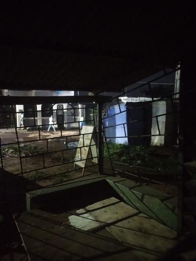 The image depicts an outdoor scene at night, with the lighting suggesting artificial illumination from the right. In the foreground, a dark, metallic fence structure obscures the view of the scene beyond. Through the fence, on the left side of the image, a paved area is visible with a few people walking. One person, dressed in white, is walking away from the viewer towards the center of the frame. Further to the left, another person in white is also walking. The background shows buildings with decorative metalwork on the facade, and some areas are illuminated, casting shadows. To the right of the frame, an A-frame sign with text is partially visible, leaning against a structure. Behind the A-frame sign, a blue structure and a white structure are visible, possibly temporary shelters or buildings. The ground appears to be wet in places, indicating recent rain or a damp surface. The overall impression is a dimly lit urban or semi-urban environment, possibly a commercial area or residential street in Agbara, Nigeria.