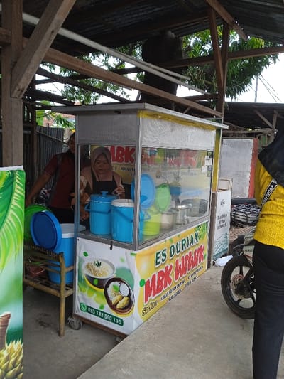 The image captures an outdoor food stall operating under a rudimentary wooden and corrugated metal shelter in Sarimulyo, Indonesia. At the center, a silver-framed glass display cart, advertising "ES DURIAN MBK LULUK" with Indonesian text, food images (including durian dishes, noodles, and rice), and a phone number (083 143 860 136), occupies the foreground. A female vendor, wearing a light-colored hijab, a patterned beige-and-brown top, and a black apron, stands behind the counter, actively engaged with two blue lidded containers and other metal bowls inside the cart. To her left-rear, a male in a dark red patterned short-sleeved shirt is partially visible. On the far right, the lower body of a person wearing a yellow long-sleeved shirt with a "Louis Vuitton" branded strap and black pants stands next to a black Honda motorcycle, of which only the front wheel and engine section are visible. Adjacent to the stall, a blue cooler or large lidded container rests on a low wooden stand. The ground is concrete, and the background includes a corrugated metal fence, green foliage, and a partial white sign with Indonesian text.