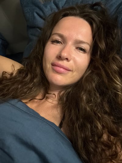 A woman with long, curly brown hair is lying down and looking at the camera. She is wearing a blue top and a delicate gold necklace. Her skin appears dewy and natural. The background is dark and blurry, with a hint of blue fabric, possibly a blanket or pillow. The lighting is soft, creating a relaxed and intimate atmosphere. There is no visible indication of the location being in the city of Изумруд Снт, Russia, or any other specific urban or rural setting. The focus of the image is solely on the woman and her direct gaze.