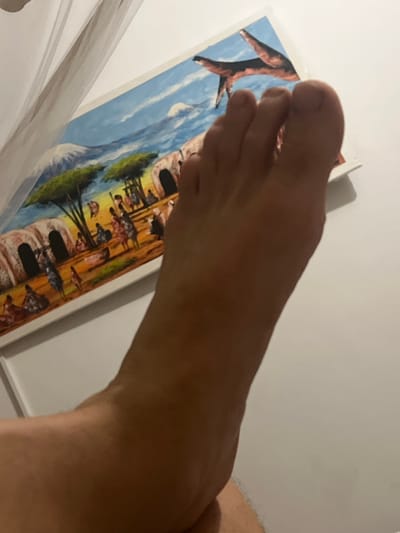 The image shows a close-up view of a human foot in the foreground, positioned over a painting in the background. The painting depicts a scene with traditional African huts, figures of people, trees, and mountains under a blue sky with clouds. The painting appears to be a landscape of a rural African village, potentially Paje, Tanzania, given the context of the request, which often features such artwork reflecting local life and scenery. The foot is dominant in the frame, obscuring a portion of the painting. The background is a plain white surface, suggesting an indoor setting. The lighting is soft, casting subtle shadows. There are no other discernible people or objects in the immediate vicinity.