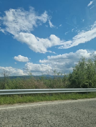 A roadside scene, possibly in or near Брусовeте, Bulgaria, shows a view of a blue sky with white clouds, roadside vegetation, a metal guardrail, and a paved road. The sky dominates the left side of the image, while the road and adjacent vegetation occupy the right side. The guardrail runs vertically between the vegetation and the road.