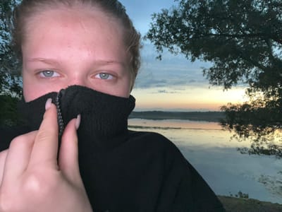 A young female with light-colored hair is partially visible in the foreground of the image. She is wearing a black fleece garment, with the collar zipped up high, obscuring her mouth and chin. Her eyes, which are light blue, are visible above the collar, and she is holding the zipper tab with her right hand. The background depicts a serene natural landscape at dusk. A body of water, likely a lake or river, reflects the soft colors of the sky, which is a gradient of blues, pinks, and yellows. Silhouetted trees are visible on the right side of the frame, and a distant treeline borders the far side of the water. The overall atmosphere is peaceful and tranquil. The image was taken in the city of Klepiki, Russia.