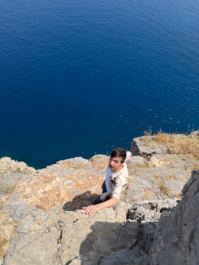A man is shown from an overhead angle, clinging to the edge of a rocky cliff face in Lindos, Greece. He is wearing a light-colored shirt and jeans, his hand gripping the rock. The blue water of the Aegean Sea stretches out below and to the side, contrasting with the grey and brown rock. The cliff edge is sparsely covered with vegetation.