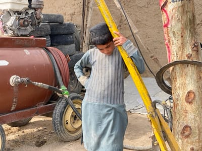 A young boy, wearing a gray and white striped shirt and blue pants, leans against a yellow metal pole. He is standing in front of a brown water tank on wheels with an attached hose, and a pile of stacked tires. The scene is in Panjwai, Afghanistan, a district in Kandahar Province, located at coordinates 31.542 latitude and 65.463 longitude. The background shows a dirt road, a tree with a thick trunk, and a mud brick wall. The boy looks to be between 10 and 12 years old. There is no one else present. The pole and water tank appear to be part of a construction or irrigation project.