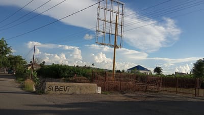 The image depicts a roadside scene in Geulangganggampong, Indonesia, under a partly cloudy sky. The foreground shows a paved road with a concrete barrier on the right side, which is graffitied with the words "SI BEUT". Behind the barrier, a rustic wooden fence encloses an area with dry vegetation. Further back, a tall, empty billboard frame stands prominently. Beyond the fence, a modern building with a blue roof is visible, surrounded by trees and other structures. The background is dominated by a vast expanse of sky filled with dramatic cumulus clouds and power lines crisscrossing the frame. A few trees line the left side of the road, and a small vehicle is seen in the distance. The overall impression is one of a developing or semi-urban area with elements of both natural landscape and man-made structures.