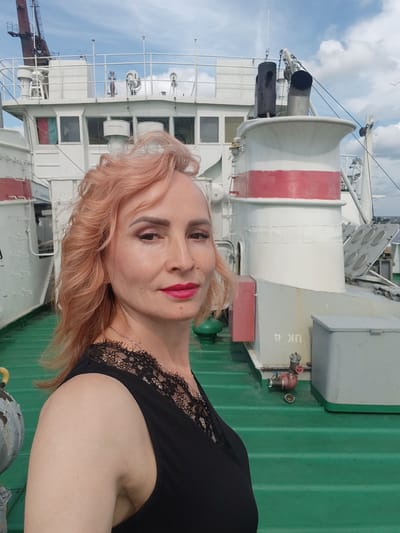 A medium shot shows a woman with wavy, peachy-pink hair and bold pink lipstick standing on the deck of a ship. She is wearing a black sleeveless top with lace detailing around the neckline. Behind her, the white superstructure of the ship is visible, including windows, railings, and large exhaust pipes. The deck is painted a vibrant green. The sky above is blue with scattered clouds. The image is taken in Faliukovo, Russia.
