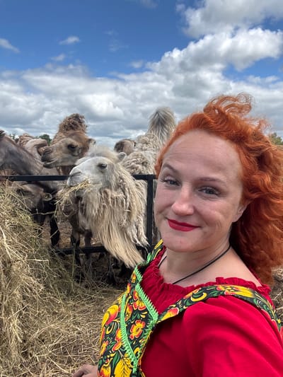A woman with bright red, curly hair smiles for a selfie as several camels eat hay behind her. She wears a red dress with a black and floral print trim. The camels are a mix of brown and light tan and appear to be in an outdoor enclosure with a fence and scattered hay. The sky is blue with white clouds. The scene is likely in Tri Ozera, Russia, given the context of the location.