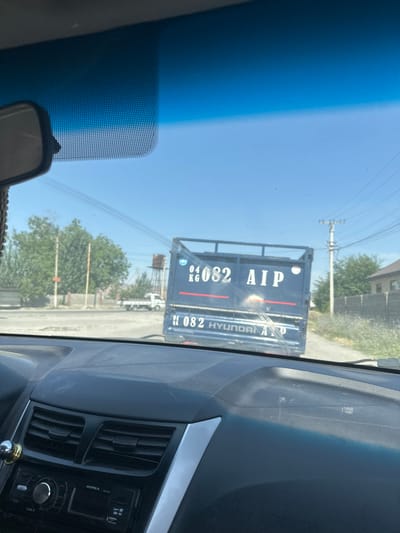 This is a first-person perspective media file, likely taken from inside a vehicle, looking forward through the windshield. The primary subject is a dark-colored truck with "AIP" written in large white letters on its side, along with the number "04 082". Below this, the words "HYUNDAI ALID" are also visible. The truck appears to be in motion or parked on a road, with other vehicles and a building visible in the background.

The setting appears to be outdoors, during the daytime, under a clear blue sky. The presence of trees and a somewhat dusty road suggests a rural or semi-urban environment. The time of day is likely mid-morning to late afternoon, given the bright sunlight. There are no people clearly visible within the frame.

Notable details include the condition of the road, which seems to be paved but with some gravel. Power lines and a utility pole are visible against the sky, indicating infrastructure in the area. A small rearview mirror of the vehicle from which the media is being captured is visible in the bottom left corner. The dashboard of the vehicle, including air vents and a radio, is also visible on the right side of the frame. The overall impression is a snapshot of daily life and travel in a location with a mix of natural and man-made elements. The visible text on the truck, "AIP 04 082 HYUNDAI ALID," is a significant detail, likely indicating identification or ownership of the vehicle. Given the location context of Ынтымак, Kyrgyzstan, this could be a local delivery or commercial truck.