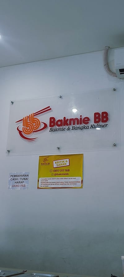 The image shows a close-up of a sign for "Bakmie BB," a restaurant. The sign is a clear acrylic plaque with the restaurant's logo and name in red and orange lettering. Below the main sign, there is a yellow sign with a list of customer service information, including a phone number and social media handle. There is also a smaller sign on the left that says "PEMBAYARAN CASH / TUNAI HARAP UANG PAS." The background is a plain white wall. The image is taken from a slightly low angle, looking up at the sign. There are no people visible in the image. The overall impression is of a clean and professional establishment. The specific location of Medang, Indonesia, is not ascertainable from the image itself.