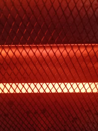 The image shows a close-up of a red-hot electric heater element behind a protective metal grid with a diamond pattern. The heating element glows brightly in the center, creating strong shadows of the grid on the surrounding surfaces. The overall color is a deep red-orange, indicative of intense heat. The location in Villa Ballester Noroeste, Argentina, is not discernible from the image.