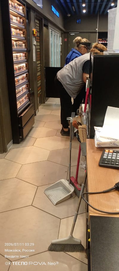 The image depicts the interior of a store, likely a convenience store or a kiosk, located in Mozhaysk, Russia. In the foreground, a broom and dustpan are placed on a hexagonal tiled floor, suggesting recent cleaning activities. To the right, a wooden counter is visible with a calculator and a stack of paper products. Behind the counter, two individuals are partially visible. One person, wearing a grey vest over a blue jacket, is bending down. Another individual, also with hair tied up, is in the background. A display rack filled with cigarette packs is prominent on the left side of the image. The overall environment appears to be utilitarian and functional, with various store fixtures and merchandise. The date stamp indicates the image was taken on July 11, 2026, at 03:29 AM.