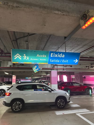 This image captures an indoor parking garage in Ondara, Spain, evidenced by the parking signs in Catalan, Spanish and English. The signs point towards the exit and access. The garage has multiple levels, indicated by the color-coded columns. Cars are parked on the right side of the image, including a white SUV and a red car. Overhead lighting fixtures and pipes run along the ceiling. A red, circular light fixture is visible on the left. The perspective is looking into the parking garage, highlighting its interior structure and signage.