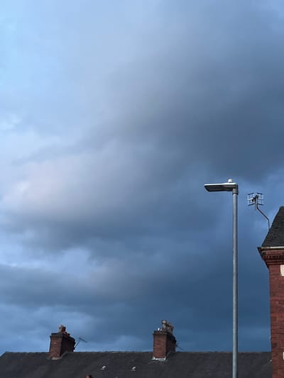 This image captures a cloudy sky over a suburban area in Stoke-on-Trent, United Kingdom. The sky is dominated by dark, heavy clouds, suggesting impending rain or a storm. In the foreground, the dark rooftops of houses are visible, with two brick chimneys protruding from one roof. A streetlamp with a modern, rectangular light fixture stands tall on the right side of the frame, its metallic pole contrasting with the dark sky. Attached to the streetlamp's pole is a small aerial antenna. To the right of the streetlamp, the corner of a red brick building with white decorative trim is visible. The overall impression is of a somber, overcast day in an urban residential setting.