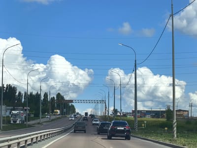 The image depicts a sunny day with scattered white clouds in a bright blue sky, viewed from a moving vehicle on a highway. The highway is flanked by green grass and trees on the right and a row of buildings and trees on the left. Several cars and trucks are visible on the road, indicating moderate traffic. Streetlights are present along the highway, and power lines are strung between poles. In the distance, there appears to be an industrial or commercial area with buildings and signage. The location is identified as Зоринский, Russia.