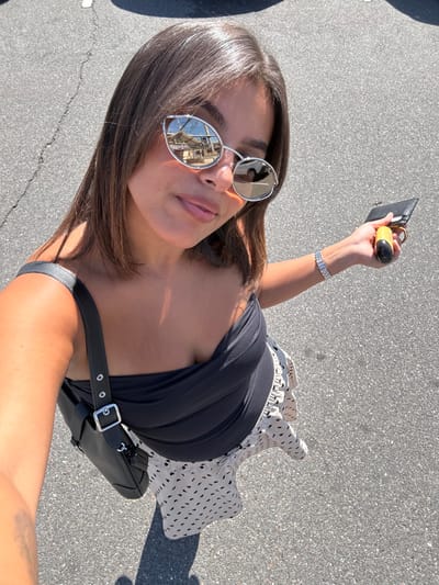 A female subject is depicted in a high-angle selfie, standing on asphalt pavement in West Windsor, United States, under bright sunlight. She has brown, shoulder-length hair and wears silver-framed, mirrored sunglasses, a black strapless top, and a light-colored skirt with dark, irregular dashed patterns. A black shoulder bag with a silver buckle strap hangs on her left side. On her right wrist, she wears a silver-toned watch or bracelet. Her right hand holds a black wallet or card holder and a yellow-capped cylindrical object. Reflections in her sunglasses reveal an outdoor structure, possibly a canopy or awning, in the left lens, and a white vehicle in the right lens. Shadows are visible on the pavement.