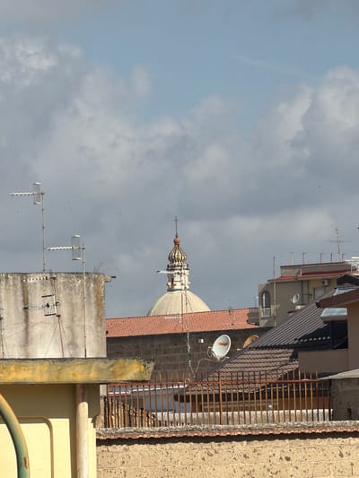 The image captures an urban landscape view from an elevated vantage point in Maddaloni, Italy, under a cloudy sky. The upper left portion of the frame shows a pale blue sky with scattered grey and white clouds.
The most prominent subject is a distinctive dome and spire of a religious building, likely a church, situated in the mid-ground. The dome is covered in reddish-brown tiles, and the spire features intricate, possibly mosaic or painted, patterns in yellow, orange, and dark colors, topped with an ornate finial.
Surrounding this landmark are various rooftops and upper levels of buildings. Several structures have traditional reddish-brown terracotta tile roofs, while others are flat concrete. One building displays a brown corrugated metal roof. Numerous television antennae and at least two white satellite dishes are mounted on the rooftops, indicating residential use.
In the foreground and towards the right, the textured, light brown stone or concrete wall of a building is visible, capped by a long, rusty metal railing composed of vertical bars. Other building facades are light-colored (beige/yellow) with dark window frames. Concrete surfaces exhibit weathering and discoloration.
Small black specks, likely birds, are seen flying in the sky, especially around the dome and above the roofs. The lighting suggests it is daytime, possibly late morning or afternoon, with soft illumination despite the clouds. There is no specific activity or event taking place, presenting a static urban scene. No visible text is present.