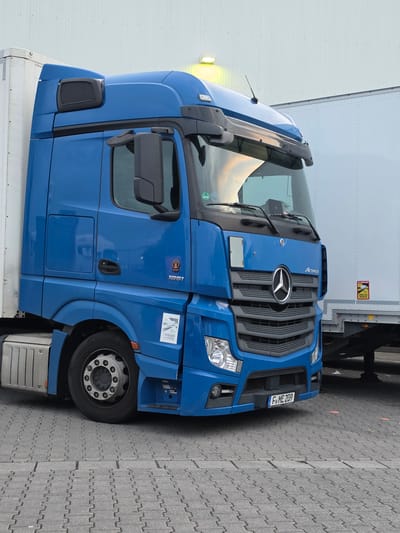 A blue Mercedes-Benz Actros semi-truck is parked on a grey, interlocking paver surface in front of a white building. The truck has a silver grille with the Mercedes-Benz star logo. A license plate on the front of the truck reads "F-ME 209". A sticker on the side of the truck reads "ANGLES MORTS" in black lettering. The truck is positioned in Zeppelinheim, Germany.