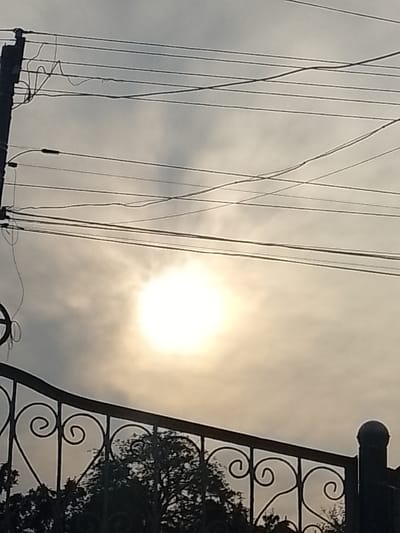 This image captures an outdoor scene in Vulcănești, Moldova, under a cloudy sky with the sun shining through, creating a bright, diffused light. A utility pole with numerous electrical wires is visible on the left, stretching across the frame. Below the wires and the sun, a decorative black metal fence with scrollwork is in the foreground. Behind the fence, silhouetted trees are visible. The overall impression is of a somewhat hazy, overcast day, with man-made infrastructure and natural elements present.
