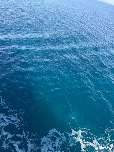 The image displays a body of water with a gentle ripple. The water transitions from a lighter blue in the upper portion to a deeper, richer blue towards the bottom left. White foam is visible in the lower left corner, suggesting movement or disturbance in the water. The scene is devoid of any people, structures, or identifiable landmarks, making it impossible to determine a specific location within the Municipal Unit of Kastro - Kyllini, Greece. The overall impression is one of an open expanse of water, possibly the sea or a large lake.