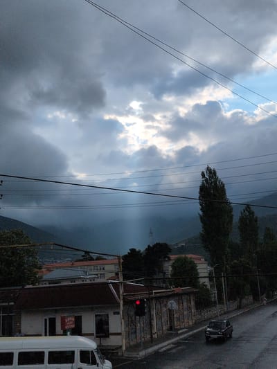 In Şəki, Azerbaijan, a dark sedan drives down a wet, asphalt road under a heavily overcast sky, illuminated by a single red traffic light. To the left, a white van is parked in front of a building with a sign that reads "SƏRIN AILƏVİ RESTORAN." The street is lined with buildings, trees, and power lines. In the background, rolling hills are visible through a light rain or mist.