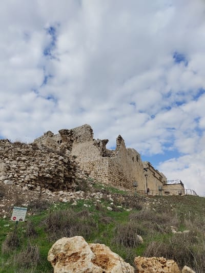The image displays the ruins of the Antipatris Fortress, located within Tel Afek National Park near Rosh HaAyin, Israel, observed from a low angle. The central subject is a large, partially collapsed stone structure constructed from light-colored, irregular blocks, situated on an elevated, rocky mound. Significant portions of the structure are in disrepair, with substantial rubble piles accumulated against its lower walls, particularly on the left side.

The foreground and midground consist of a sloping terrain covered with sparse, dry green and brown vegetation, interspersed with numerous light-colored rocks and boulders. Three prominent, tan-colored boulders are visible in the immediate foreground. On the lower left, a rectangular white sign with dark Hebrew text and a red 'no entry' symbol is present. To the right of the main ruin, a more preserved or reconstructed section of a building is visible, featuring a dark metal railing and what appears to be a utility pole or light fixture. The sky above is partially covered by scattered white cumulus clouds against a blue background. No individuals are visible in the scene.