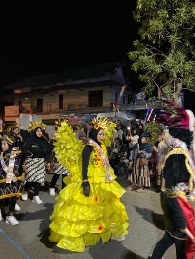 A nighttime street procession in Kedungbanteng, Indonesia, is captured near an archway sign reading "DESA KALIDAN". The central figure, a female, is positioned slightly right of center, walking towards the right. She wears a voluminous, multi-tiered gown constructed from bright yellow material, a black long-sleeved top, dark sheer gloves, and a black hijab. Her head is adorned with a golden, radiating headdress featuring sunflowers. A sash, decorated with sunflowers and Indonesian rupiah banknotes, drapes from her left shoulder. Behind her left shoulder, another individual wears a matching yellow, tiered backpiece. To the far left, a person in black and gold traditional attire with a banknote-laden sash and headdress walks right-to-left. A woman in a black hijab and a black-and-white patterned batik skirt is visible between these figures. On the extreme right, part of a distinct feathered costume, likely a headdress with pink, black, and white elements, is visible. The street is populated by numerous spectators, many utilizing mobile phones to record the event. In the background, multi-story buildings with dark roofs are illuminated by ambient light. A prominent green-leafed tree stands on the right. An Indonesian flag is partially visible on the left. The ground is dark pavement with a faint blue line on the left.