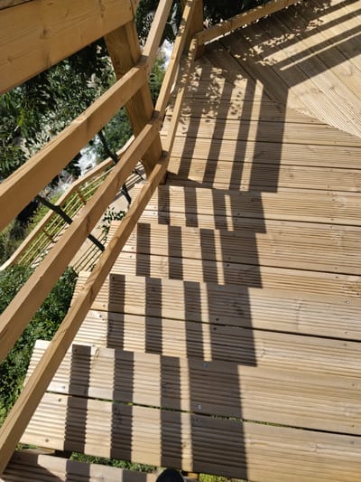 The image shows a wooden deck or platform in Alijó, Portugal. The deck is constructed of grooved wooden planks and is surrounded by a wooden railing. The railing is partially visible, showing both vertical and horizontal wooden bars. Shadows of trees or foliage fall across the deck. Through the railings, one can see green vegetation. A portion of a shoe is visible at the bottom of the image.