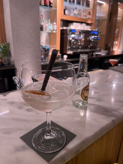 A high-angle shot focuses on a cocktail with ice and a straw in a clear glass. Next to the glass, on a marble surface, is an empty bottle of Thomas Henry Tonic Water. The drink sits on a square gray coaster. In the blurred background, a bar display features illuminated shelves with bottles and glassware. The lighting is dim, creating a relaxed atmosphere.