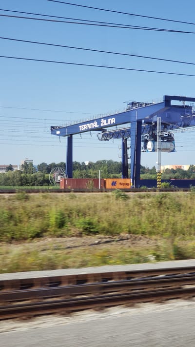 This is a wide shot of a shipping terminal in Teplička nad Váhom, Slovakia, on a clear day. The main subject is a large blue gantry crane with the words "TERMINÁL ŽILINA" written on its side. The crane spans multiple railway tracks and is positioned over a collection of shipping containers. To the left of the crane, a large, spoked industrial reel is visible, and to the right, a yellow and black striped pole stands. In the background, a line of trees and some buildings can be seen. The foreground shows blurred railway tracks and vegetation, indicating the photo was taken from a moving vehicle.