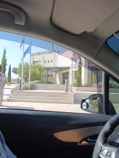 The image, taken from inside a car, shows the exterior of a light-colored, modern building with a large set of stairs leading up to its entrance. The building appears to be a hotel or resort, possibly the "Golden Mare" hotel given the partial sign visible on the left. Two flags, one Greek, fly on flagpoles in front of the building. The surrounding environment includes manicured bushes, cypress trees, and paved walkways. A side mirror of the car is visible, reflecting the environment. The sky is clear and blue. The scene is set in the Municipal Unit of Larissos, Greece.