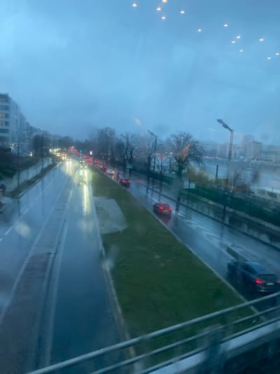 A rainy urban scene displays a wet multi-lane road with cars, partially obscured by raindrops on a window. Green median strips and bare trees line the road. Buildings are visible in the background, shrouded in mist. The sky is overcast and gray. Streetlights and car headlights are visible in the distance along the road, illuminating the wet pavement.