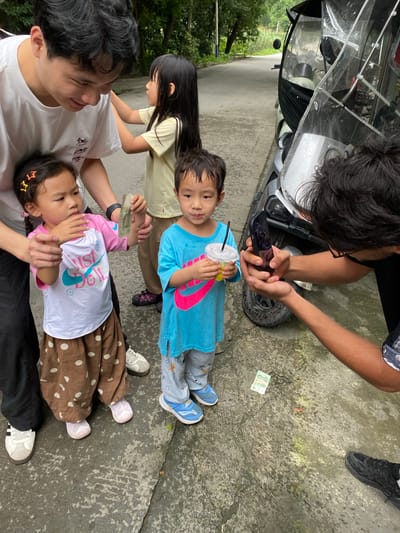 In Chongzhou City, China, on a wet, paved surface, a group of five individuals are gathered. A young boy in a blue t-shirt holds a drink with a straw. To his left, a young girl in a pink and white shirt with "Just Do It" written on it holds a folded bill. A man with black hair in a white t-shirt is looking down at the girl, with his hands near her. To the right of the boy, another individual's back is visible, holding a smartphone. Behind them, a scooter with a clear plastic covering is parked. The environment appears to be outdoors, with trees and vegetation visible in the background. The ground is damp, suggesting recent rain.