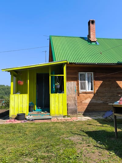 The image depicts a wooden house with a bright green metal roof and a contrasting bright yellow porch in the village of Maiskii, Russia. The house is constructed of horizontal wooden planks, with a single white-framed window visible on the right side, featuring white curtains. A red brick chimney stands on the peak of the roof. The porch is painted a vivid yellow and has vertical wooden planks. Several pairs of footwear, including sandals and closed-toe shoes, are placed on the porch steps and the adjacent ground. A black bucket and a dark green trash bin are situated on the porch. Two hanging flower baskets, one with red flowers and another with white flowers, are suspended from the porch roof. The foreground is a grassy yard with patches of bare soil. In the background, a line of trees is visible against a clear blue sky. To the right of the house, a part of a table and a folded blue chair are present. The scene suggests a residential outdoor setting on a sunny day.