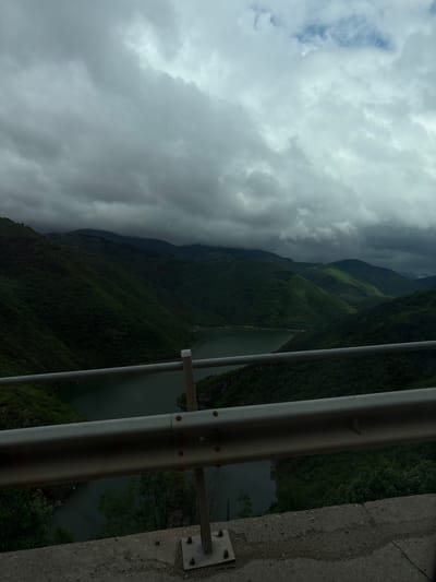 The image depicts a view from what appears to be a bridge or elevated structure near Liaskovo, Bulgaria. A series of green, forested hills slopes down to a body of water. The sky is overcast with thick, dark gray clouds dominating the upper left portion of the image. A portion of a metal guardrail is visible on the right side of the image, bolted to a concrete structure. The viewpoint suggests the presence of a man-made structure overlooking the natural landscape.