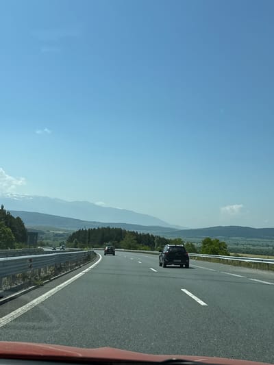 The image captures a sunny day on a multi-lane highway near Staro Selo, Bulgaria, viewed from the interior of a moving vehicle. The bright blue sky occupies the upper two-thirds of the frame, featuring a few faint, wispy white clouds. In the mid-distance, a range of mountains stretches across the horizon, with some distant peaks showing patches of snow or light rock and appearing faded due to atmospheric haze. Below the mountains, rolling hills covered in a mix of dense evergreen and deciduous forests are visible, transitioning into open, possibly cultivated, green fields in the valleys.
The asphalt highway features at least two lanes in the direction of travel, marked by white dashed lines, and a solid white line on the left shoulder. Metal guardrails line both sides of the road, separating it from the surrounding terrain. Several vehicles are present on the highway. In the immediate foreground, a black SUV (possibly a Hyundai) travels in the right lane, with its rear clearly visible. Further ahead in the left lane, a dark-colored sedan or smaller SUV is seen. Additional vehicles appear as dark specks in the distance along the highway. The bottom edge of the frame reveals a portion of a red dashboard or vehicle interior, indicating the viewer's position within a car. The overall scene suggests travel through a mountainous and rural landscape under clear weather conditions.